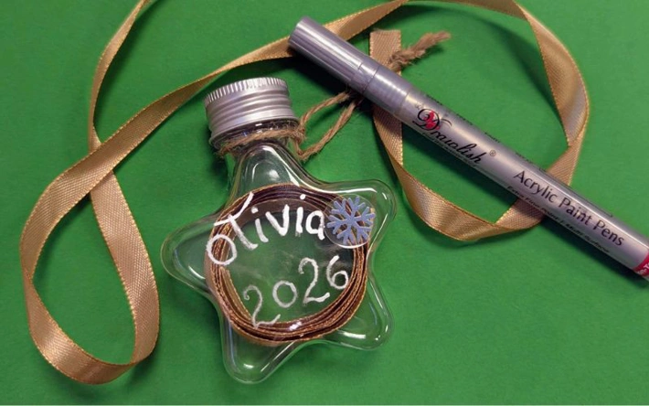 A glass bottle containing a gold ribbon, with the name 'Olivia' and '2026' written in silver paint pen, against a green background