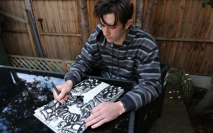 Artist Blake Walker sitting beside a garden fence and drawing in a sketch pad