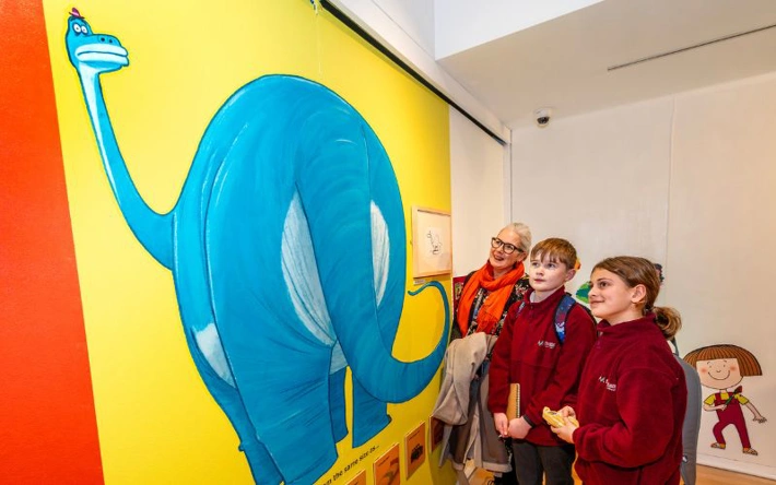 Three people looking at a large mural of a dinosaur on a wall, part of the Oi Chelmsford! exhibition. 