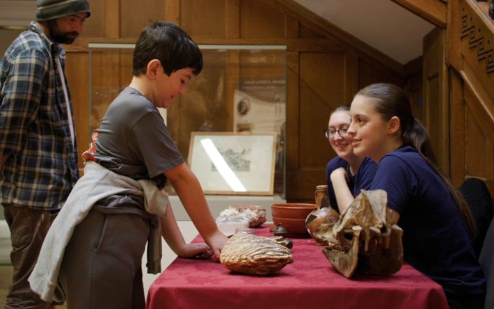A young child handles some of the objects on a red table with two museum volunteers